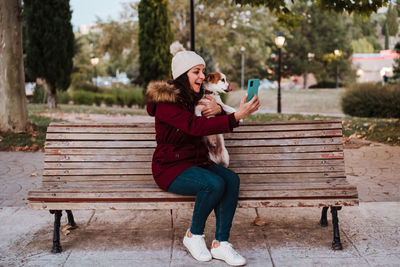 Woman with dog using phone while sitting on bench against trees