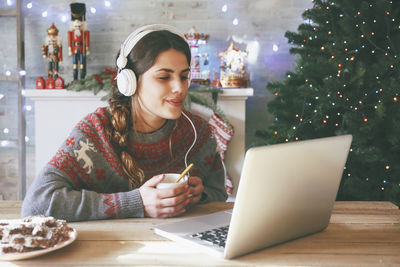 Woman with cup of coffee using laptop and headphones at christmas time