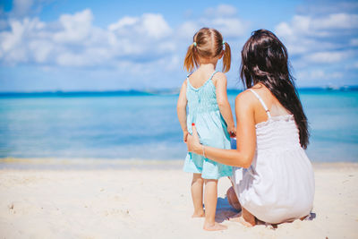 Rear view of mother and daughter crouching on sand at beach