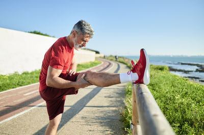 Mature man stretching leg on wooden railing by the sea during sunny day