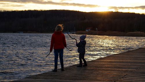 Siblings standing on pier over lake against sky during sunset