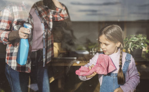 Girl cleaning window glass with rag near grandmother at home