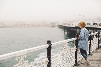 Full length of young woman standing by railing on brighton pier