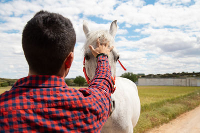 Rear view of man touching horse at ranch against sky