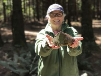 Portrait of smiling senior woman holding stone while standing in forest