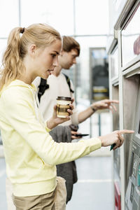 Woman using atm while holding coffee by friend at railroad station