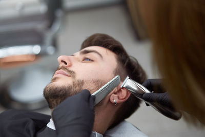 Portrait of young man who sitting in barbershop and woman hairdresser cut hair