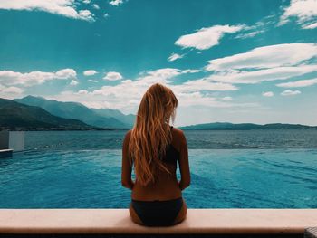 Rear view of woman sitting in bikini at poolside by sea against sky