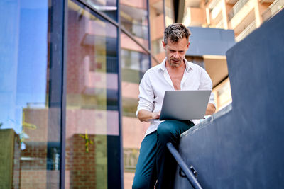 Low angle of mature businessman using laptop and smartphone while sitting on border during work on remote project on street