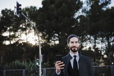 Portrait of young man holding camera while standing against trees