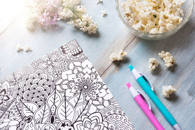 High angle view of floral pattern on paper by pens and popcorns over wooden table