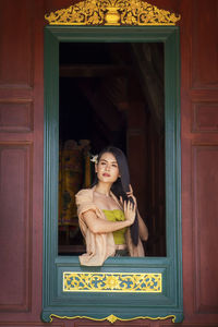  asian girl wearing thai traditional dress hand holding hair on window. looking at side.