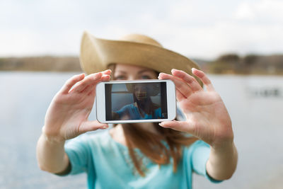 Portrait of smiling young woman taking selfie with smart phone against lake