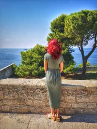 Rear view of redhead woman standing on footpath near sea against blue sky