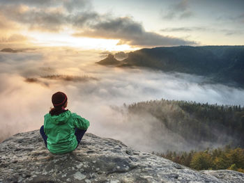 Attractive body woman sitting in green windcheater on mountain peak watching sunrise over fog