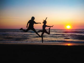 Silhouette siblings jumping on shore at beach against sky during sunset