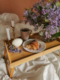 Morning coffee in bed with french croissant, candle in shell form and bunch of lilac flowers.