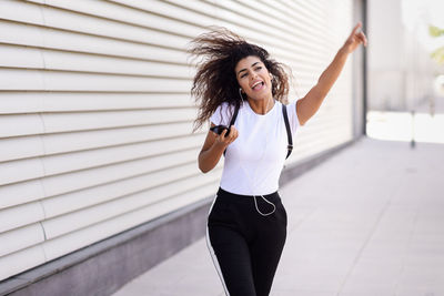 Cheerful young woman listening music through smart phone while walking on street