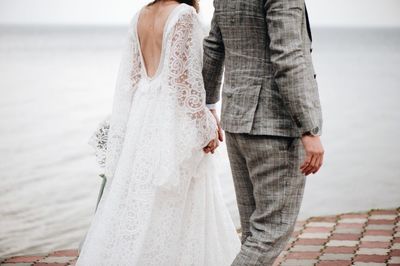 Midsection of bride and bridegroom holding hands by sea on footpath