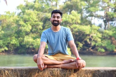 Portrait of smiling young man sitting on wall against trees