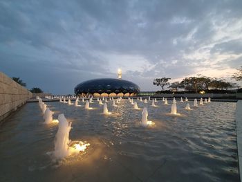 Illuminated building by sea against cloudy sky