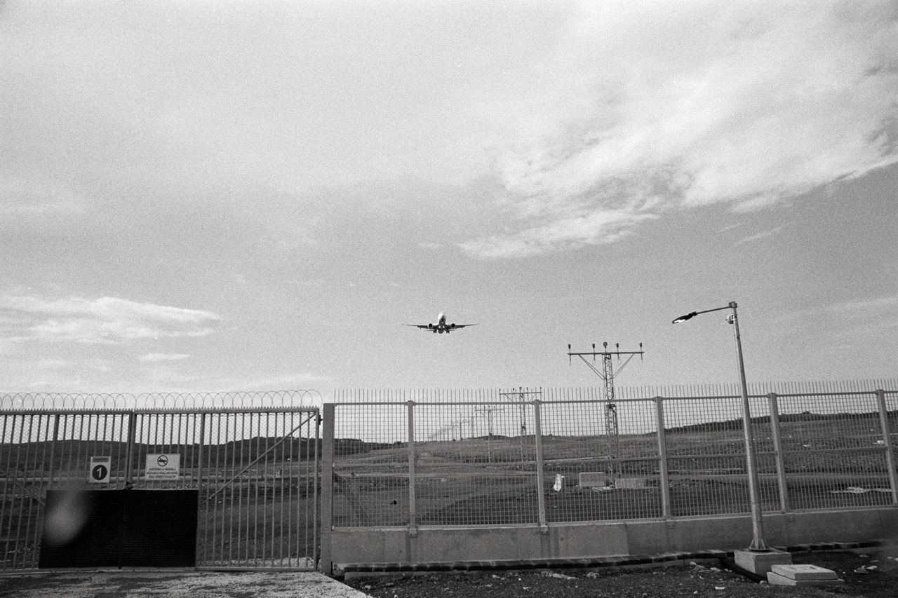 AIRPLANE FLYING OVER AIRPORT RUNWAY AGAINST SKY