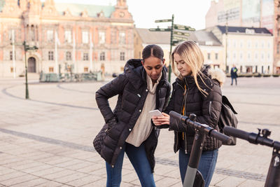 Teenage girl showing mobile phone to friend while standing by e-scooter at city