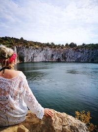 Rear view of woman sitting on rock by lake against sky