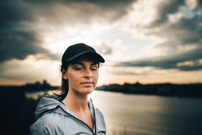 Young sportswoman with eyes closed standing on hill against cloudy sky during sunset