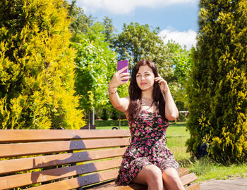 Young woman taking selfie while smart phone while sitting on bench against trees in park