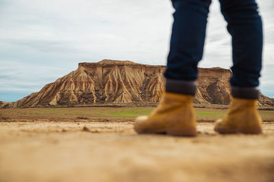 Legs on faceless traveler in brown boots and blue jeans standing on dirty sandy road with mountain and sky on blurred background in bardenas reales, navarre, spain