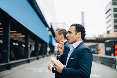 Side view of mature businessman talking through in-ear headphones while standing by male colleague in city