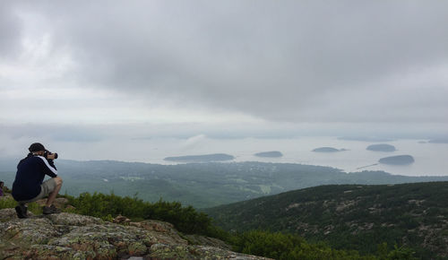 Side view of man photographing while crouching on mountain against cloudy sky