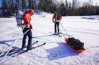 Two men attach pulk sleds to their backpacks in a snowy parking lot