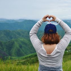 Rear view of woman with arms raised making heart shape while standing on mountain against sky