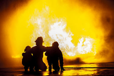 Rear view of silhouette firefighters crouching against fire at night