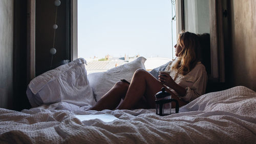 Side view of woman looking through window while sitting on bed at home