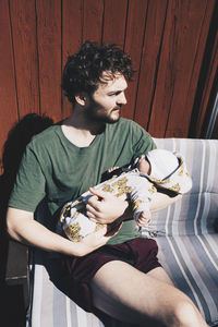 Father carrying baby boy while sitting on chair against wall during sunny day