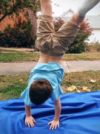 Boy practicing handstand on blue fabric at grassy field against sky