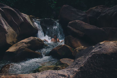 Shirtless man sitting in flowing water by rock