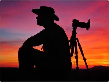 Silhouette man sitting by camera against sky during sunset