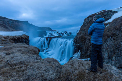 Rear view of man looking at waterfall while standing on mountain against cloudy sky