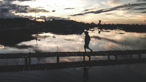 Silhouette man walking on pier over lake against sky during sunset