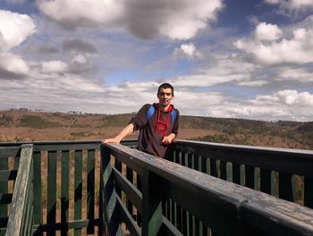Portrait of young man standing on railing against sky