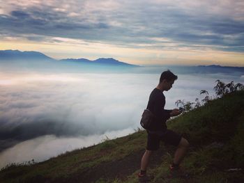 Side view of young man using smart phone while walking on mountain against cloudy sky during sunset