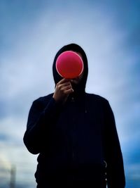 Man in black hoodie holding a red balloon in front of his face against moody cloudy blue sky.