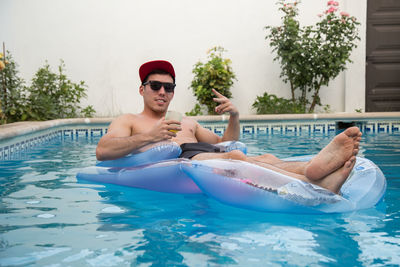 Portrait of shirtless young man with beer glass relaxing on pool raft in swimming pool
