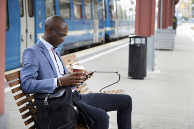 Businessman using smart phone while sitting with coffee at railroad station