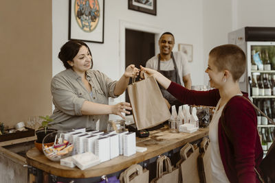 Happy female owner giving paper bag to customer at checkout counter in store