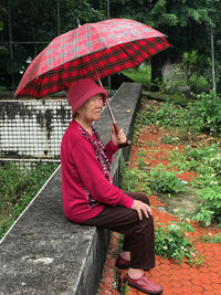 Side view of senior woman holding umbrella while sitting on retaining wall